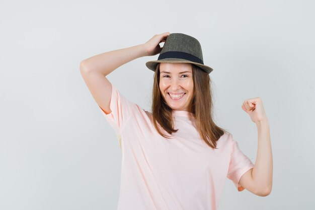 Young female in pink t-shirt, hat posing with hand on head and looking attractive , front view.