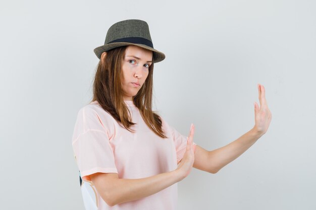 Young female in pink t-shirt, hat raising hands to defend herself and looking strict , front view.