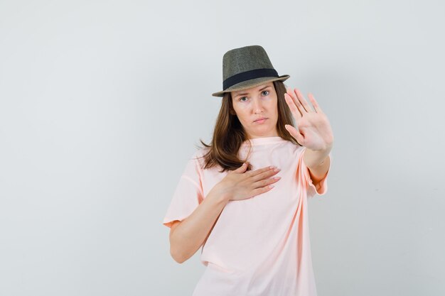 Молодая женщина в розовой футболке, шляпе, показывающей жест отказа и серьезной, вид спереди.
