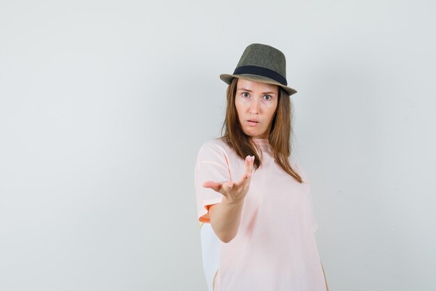 Молодая женщина в розовой футболке, шляпа, протягивая руку озадаченным жестом, вид спереди.