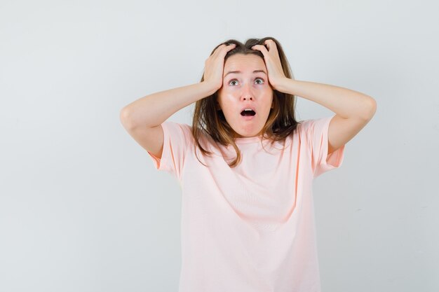 Young female in pink t-shirt holding head with hands and looking anxious, front view.