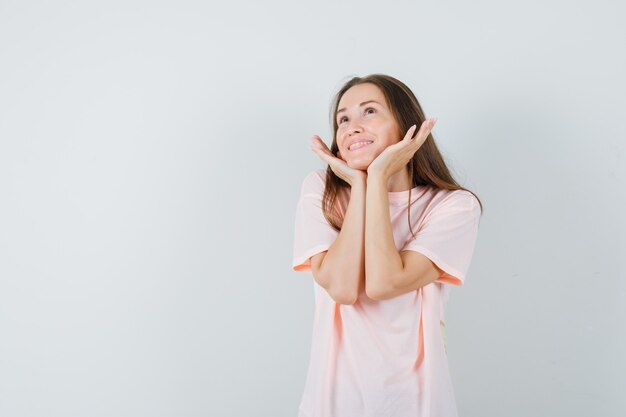Молодая женщина в розовой футболке, прикрывая лицо руками и выглядя мило, вид спереди.