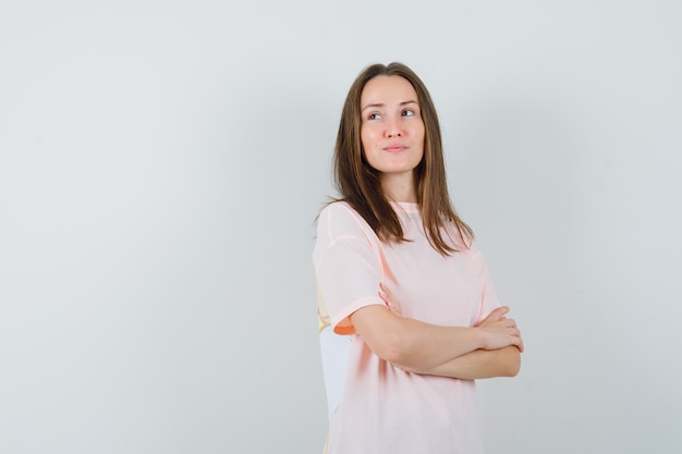 Молодая женщина в розовой футболке стоит со скрещенными руками и мечтательно смотрит, вид спереди.