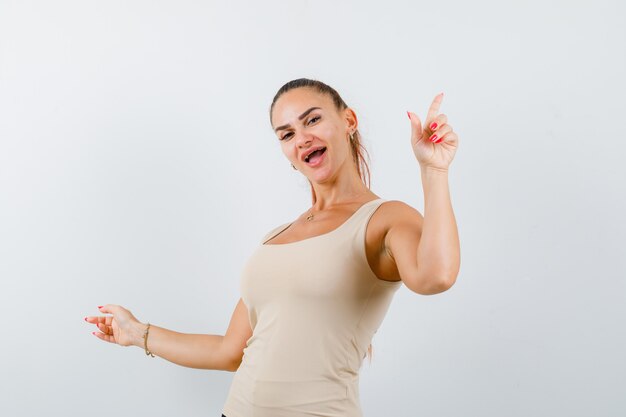 Young female pointing aside in beige tank top and looking happy. front view.