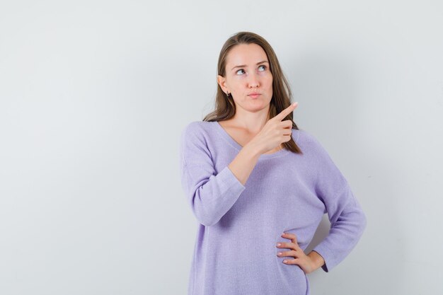 Young female pointing aside in lilac blouse and looking thoughtful 