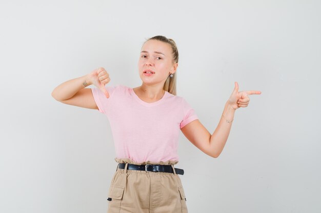 若い女性は脇を向いて、Tシャツ、ズボンで親指を下に見せて、がっかりしているように見えます。正面図。