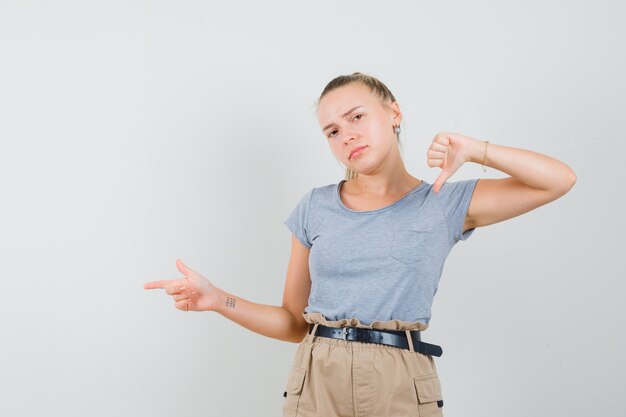 若い女性は脇を向いて、Tシャツ、ズボンで親指を下に見せ、不満を探しています。正面図。