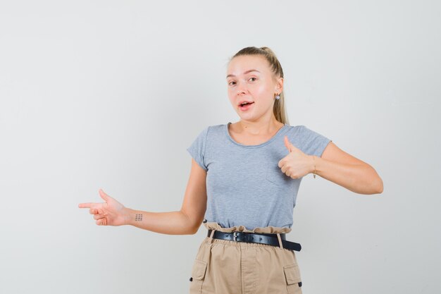 Young female pointing aside, showing thumb up in t-shirt, pants and looking happy , front view.