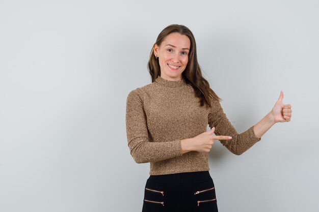 Young female pointing aside while showing thumb up in golden blouse and looking pleased. front view. space for text