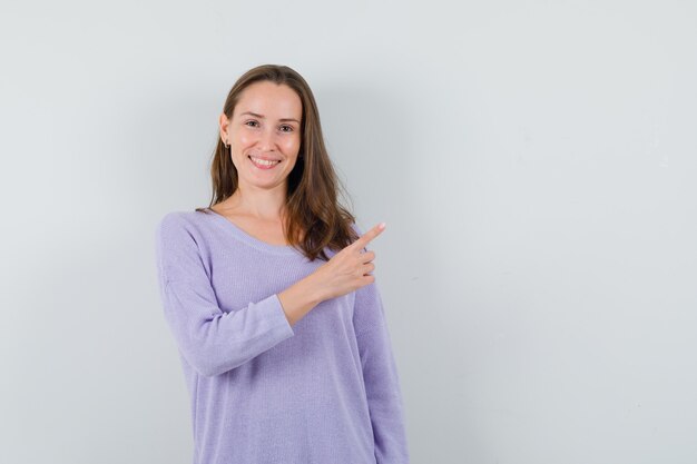 Young female pointing aside while smiling in lilac blouse and looking pleased 