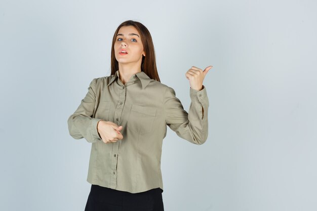 Young female pointing aside with thumbs in shirt, skirt and looking indecisive