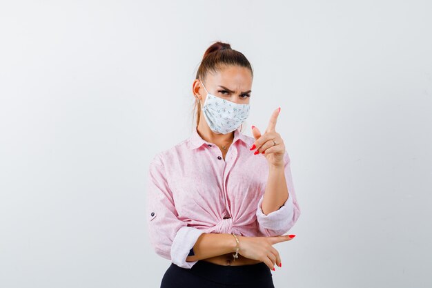 Young female pointing away in shirt, pants, medical mask and looking confident. front view.