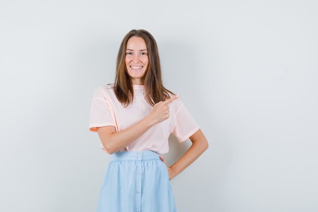 Young female pointing away in t-shirt, skirt and looking merry , front view.