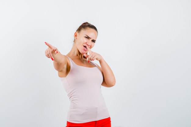 Young female pointing away in white tank top, pants and looking confident , front view.