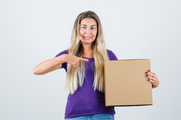 Young female pointing at box in violet t-shirt and looking attentive. front view.