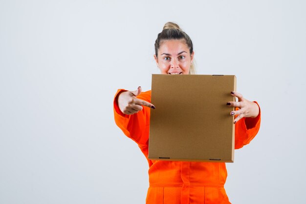 Young female pointing at box in worker uniform and looking positive. front view.