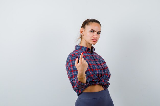 Young female pointing at camera in checkered shirt, pants and looking serious , front view.