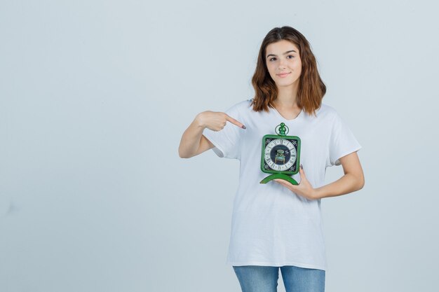 Young female pointing at clock in white t-shirt, jeans and looking cheery. front view.