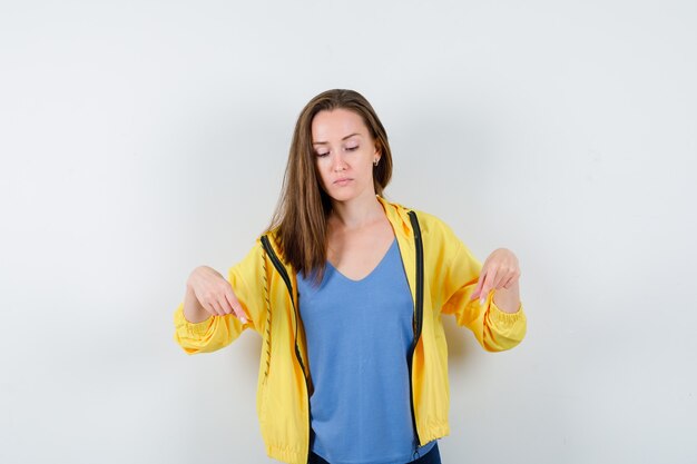 Молодая женщина указывая пальцами вниз в футболке, куртке и глядя сосредоточенно, вид спереди.