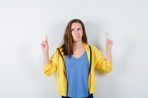 Young female pointing fingers up in t-shirt, jacket and looking hesitant. front view.