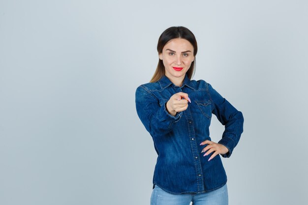 Young female pointing at front while keeping hand on hip in denim shirt and jeans and looking confident