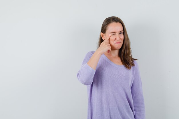 Young female pointing to her eyelid while winking in lilac blouse and looking focused 