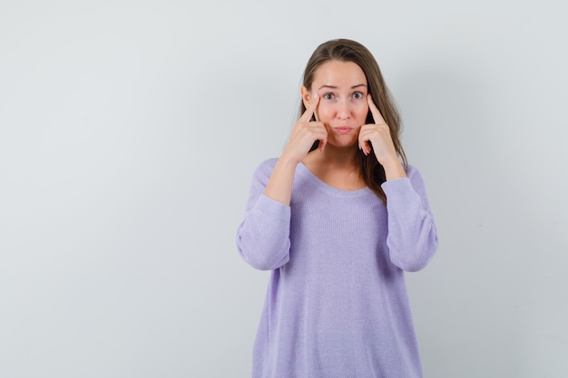 Young female pointing at her eyes in lilac blouse and looking concentrated. front view. space for text