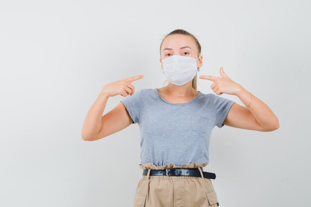 Young female pointing at her medical mask in t-shirt, pants , front view.