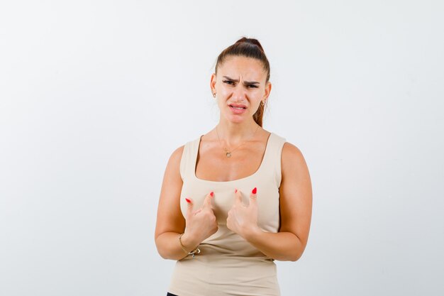 Young female pointing at herself in beige tank top and looking displeased. front view.