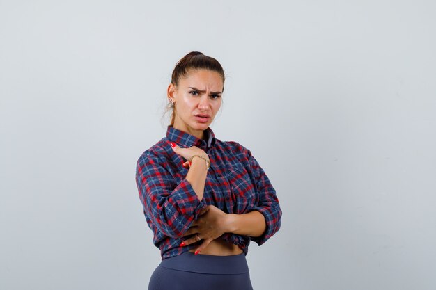 Young female pointing at herself in checkered shirt, pants and looking hesitant , front view.