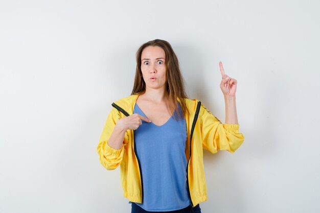 Young female pointing at herself and up in t-shirt, jacket and looking surprised, front view.