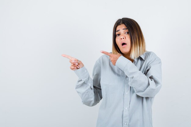 Young female pointing to the left side in oversized shirt and looking perplexed. front view.