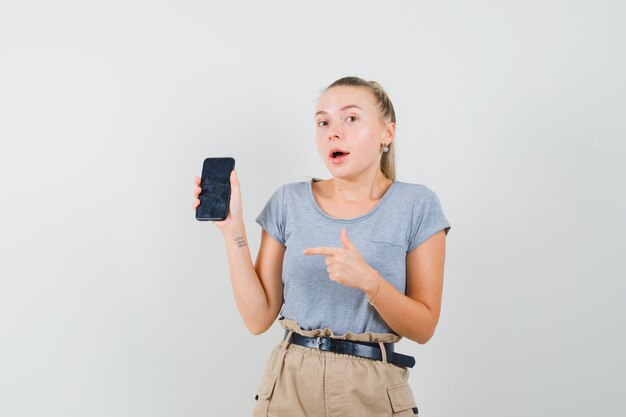 Tシャツ、ズボンで携帯電話を指差して驚いて見える若い女性、正面図。