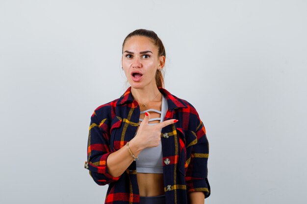 Young female pointing to the right side in crop top, checkered shirt and looking puzzled. front view.