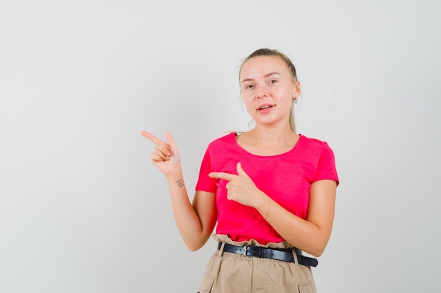 Young female pointing to the side in t-shirt, pants and looking cheerful , front view.