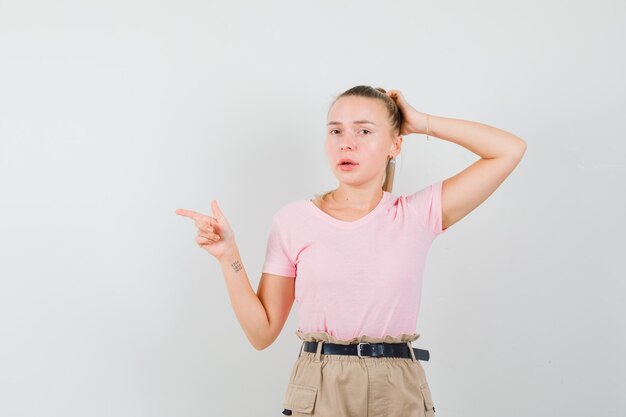 Young female pointing to the side while scratching head in t-shirt, pants front view.