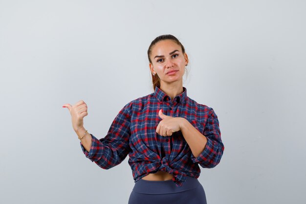 Young female pointing thumbs up to the left side in checkered shirt, pants and looking confident , front view.