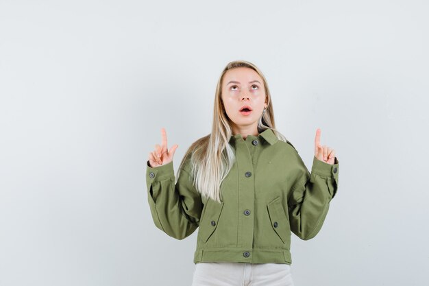 Young female pointing up in green jacket,jeans and looking concentrated. front view.