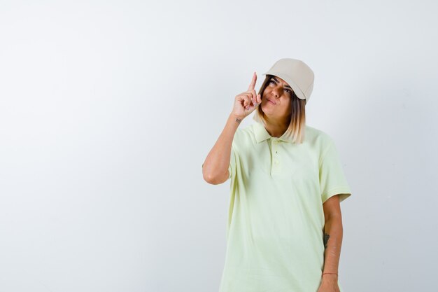 Young female pointing up in t-shirt, cap and looking hesitant , front view.