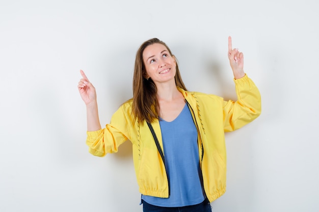 Young female pointing up in t-shirt, jacket and looking hopeful, front view.