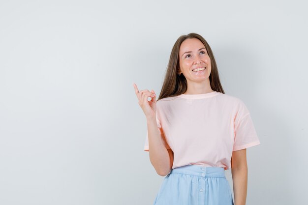 Young female pointing up in t-shirt, skirt and looking optimistic. front view.