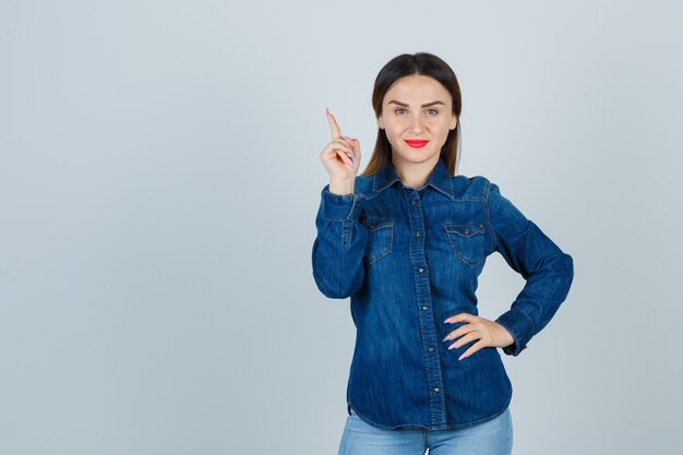Young female pointing up while keeping hand on hip in denim shirt and jeans and looking cheery