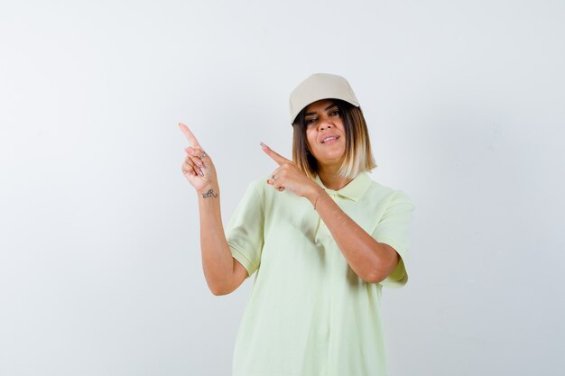 Free photo young female pointing at upper left corner in t-shirt, cap and looking cheerful , front view.