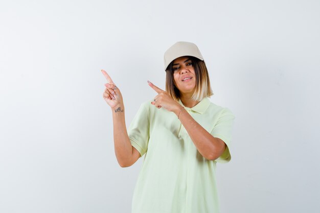Молодая женщина, указывая на верхний левый угол в футболке, кепке и выглядит веселой, вид спереди.