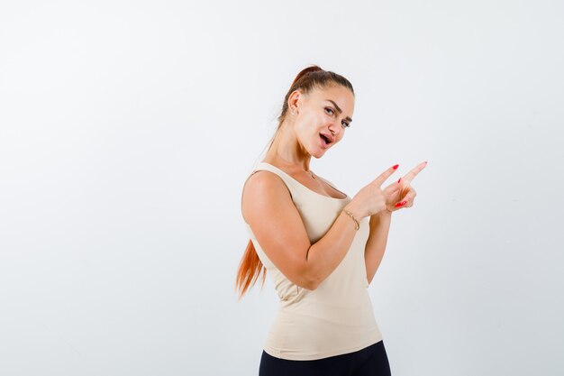 Young female pointing at upper right corner in beige tank top and looking amazed. front view.