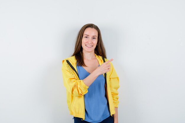 Young female pointing at upper right corner in t-shirt, jacket and looking confident. front view.