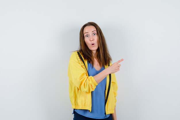 Young female pointing at upper right corner in t-shirt, jacket and looking surprised. front view.