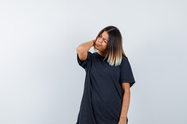 Young female in polo dress holding hand on neck and looking exhausted , front view.