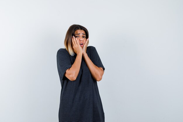 Young female in polo dress holding hands on cheeks and looking puzzled , front view.