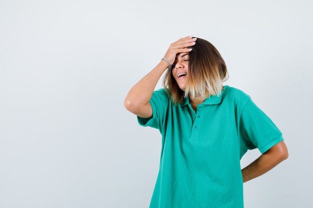 Young female in polo t-shirt with hand on forehead while keeping hand behind back and looking glad , front view.